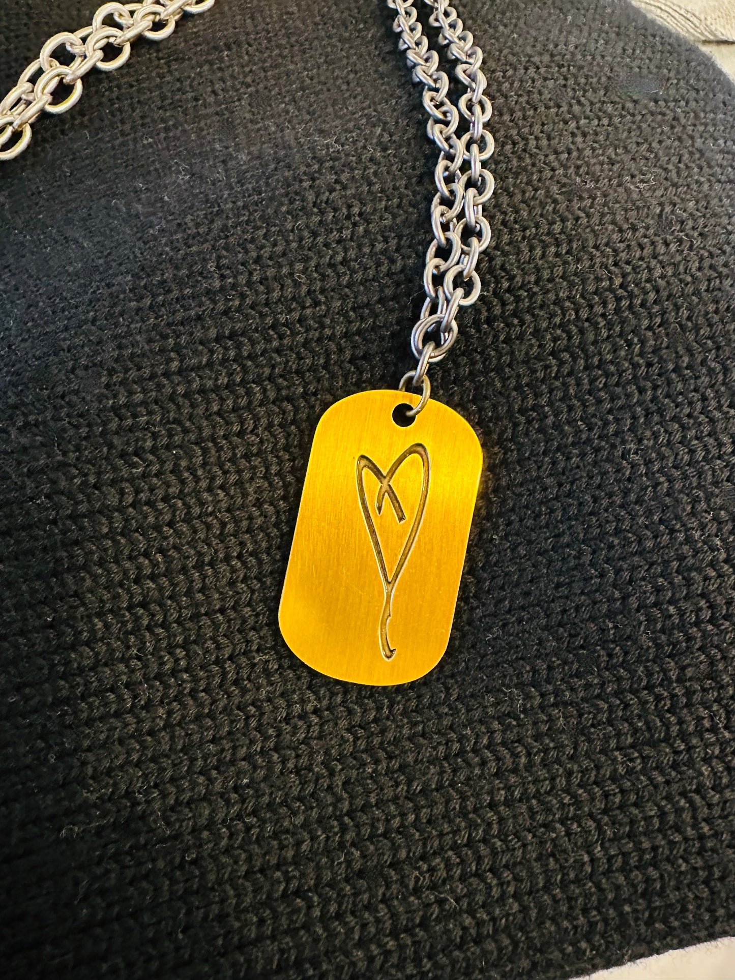 The Eartha Kitt's Hand Drawn Heart/Here's To Life - Dog Tag Necklace from My Store features a stylized gold heart pendant on a silver chain, elegantly displayed against textured black fabric, capturing the spirit of gratitude.