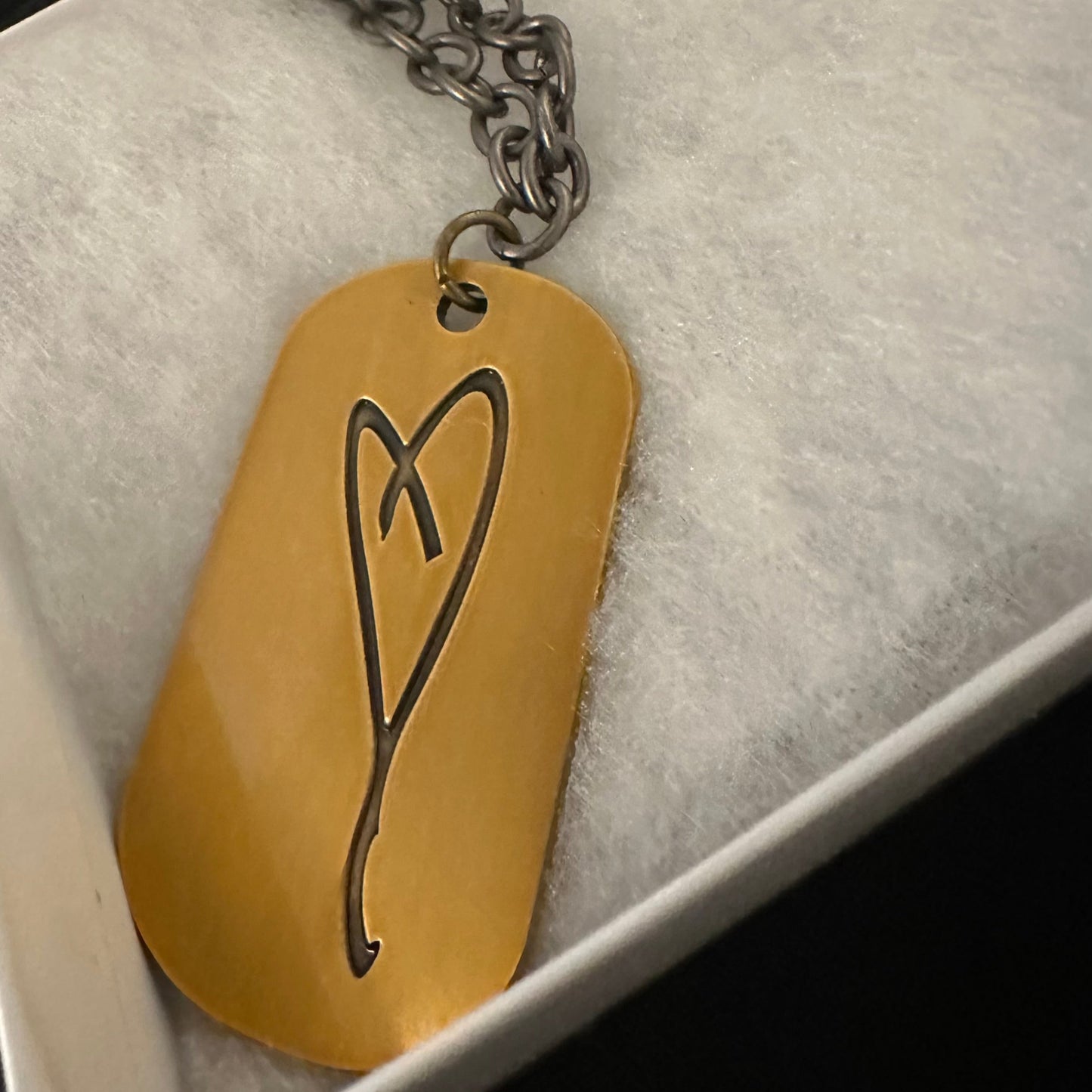 A metallic chain necklace from My Store features a gold rectangular dog tag with an engraved abstract heart, symbolizing gratitude. Named "Eartha Kitt's Hand Drawn Heart/Here's To Life," it elegantly rests on a soft, light gray surface, celebrating life.