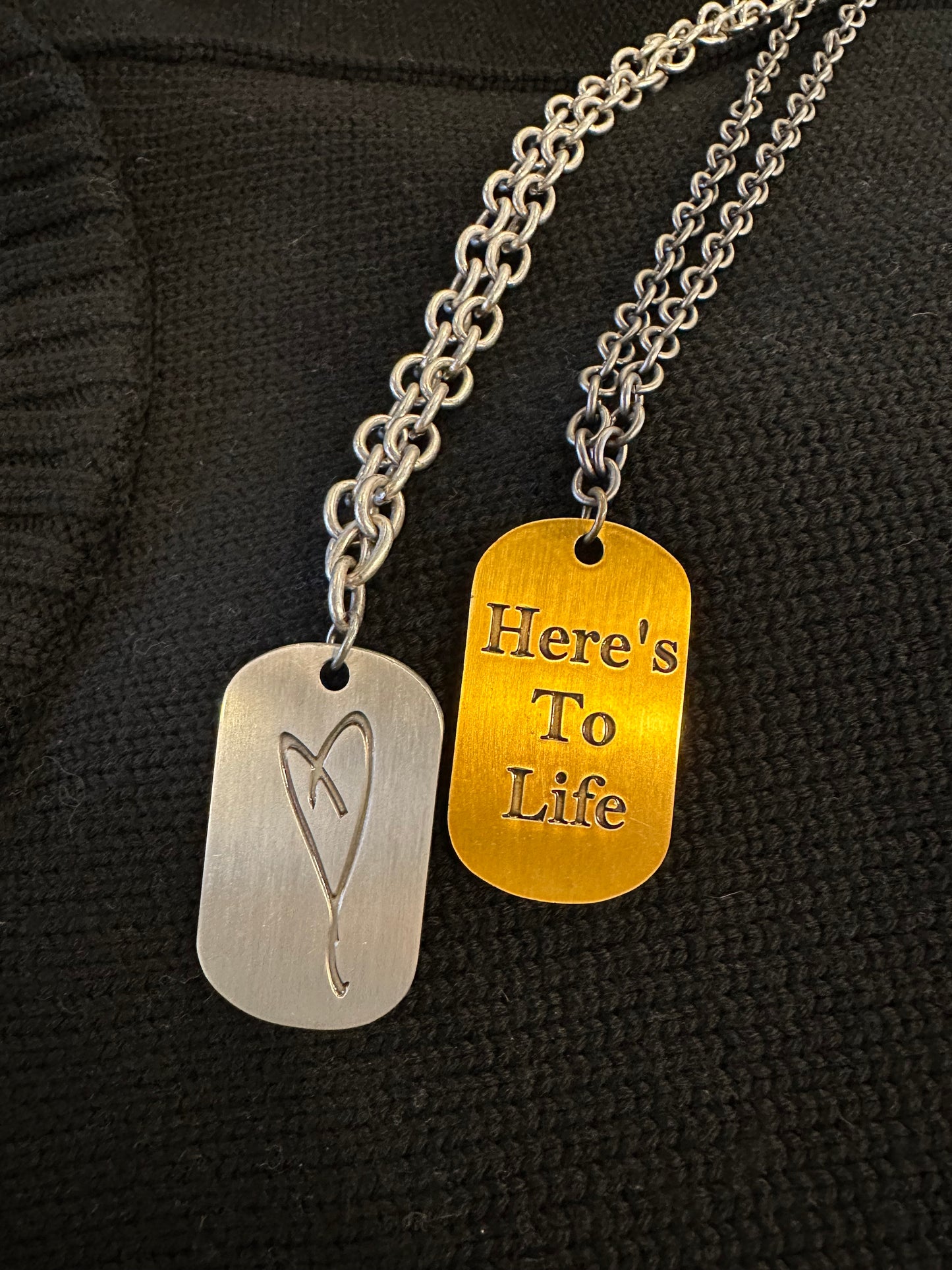 Two metal dog tags hang on a silver chain against a black background. The silver tag features Eartha Kitt's hand-drawn heart, while the gold one is engraved with "Here's To Life." A perfect testament to gratitude and joy from My Store's collection.