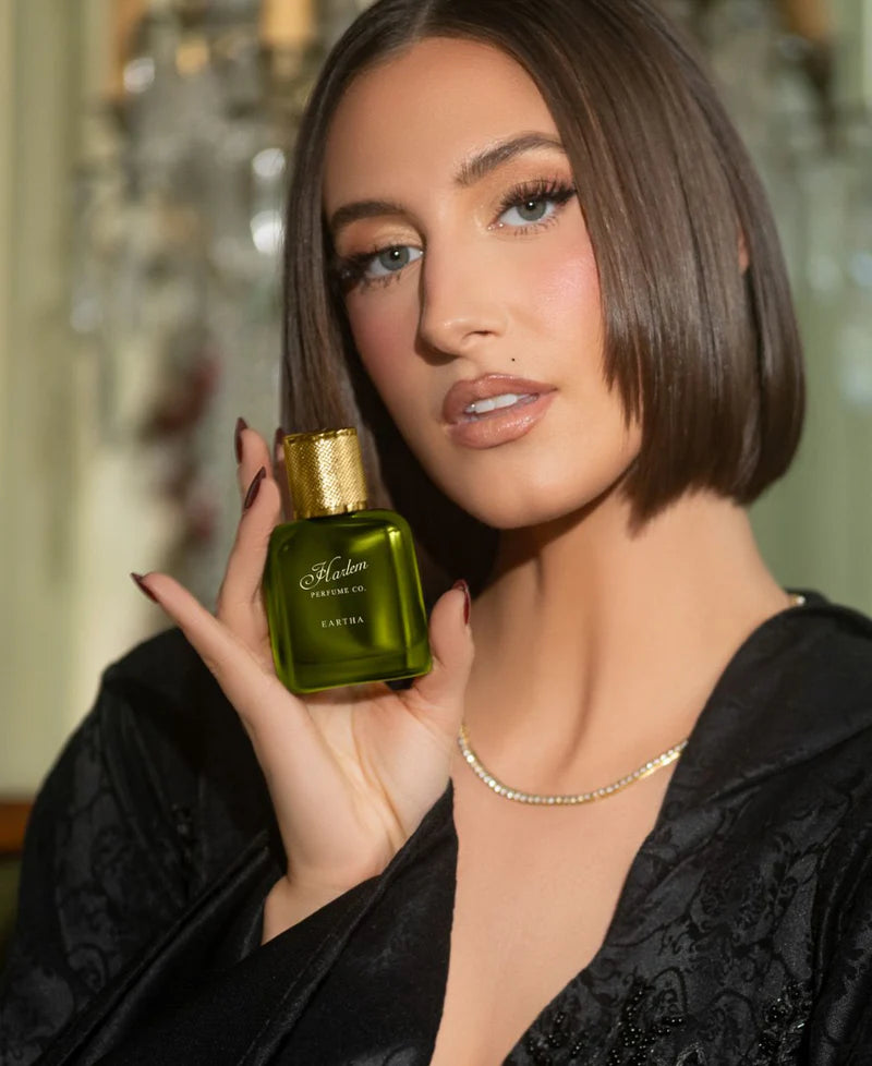 A woman with straight, shoulder-length brown hair holds up a Simply Eartha EARTHA EAU DE PARFUM bottle, featuring a green body and gold cap. She wears a black robe and gold necklace in an elegant, softly lit room.