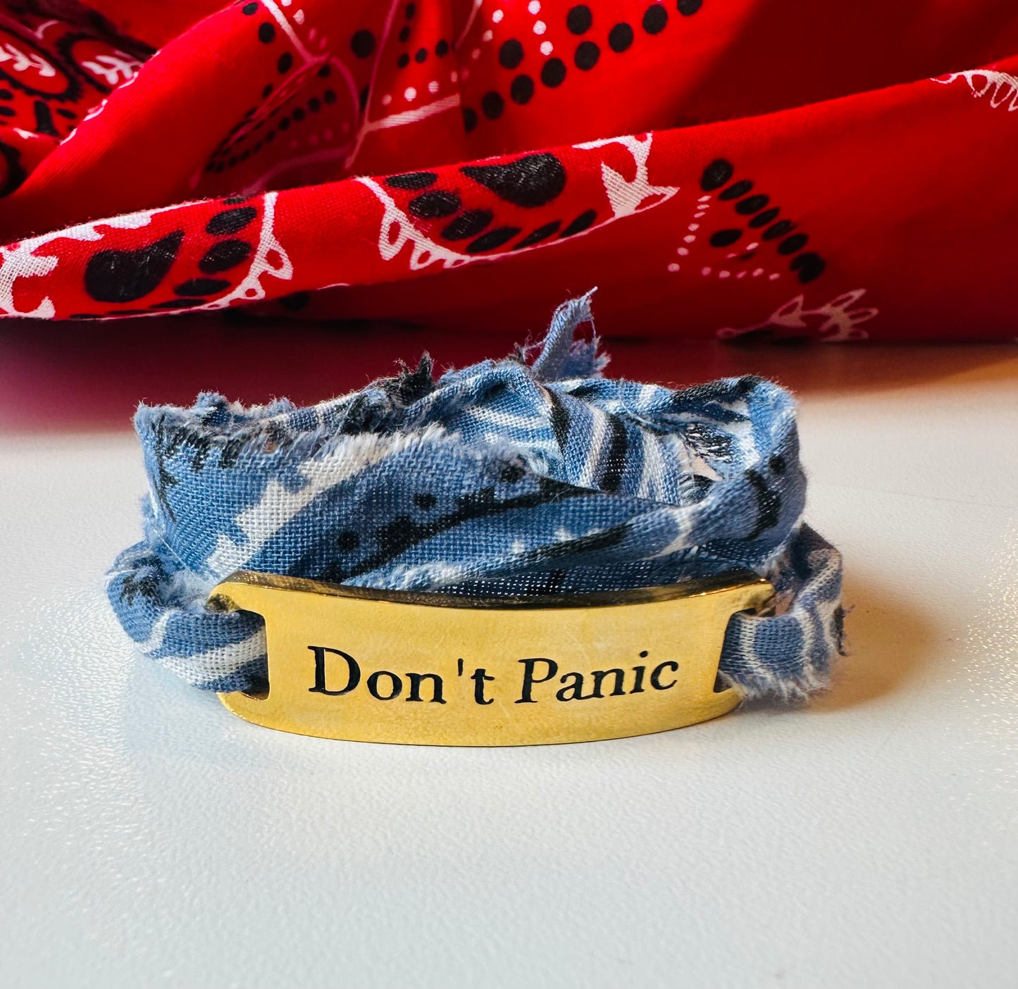 The Simply Eartha Don't Panic Bandana Wrap Bracelet features a limited gold plated design engraved with "Don't Panic," wrapped in frayed blue fabric, set against a vibrant backdrop of red cloth with black and white patterns.