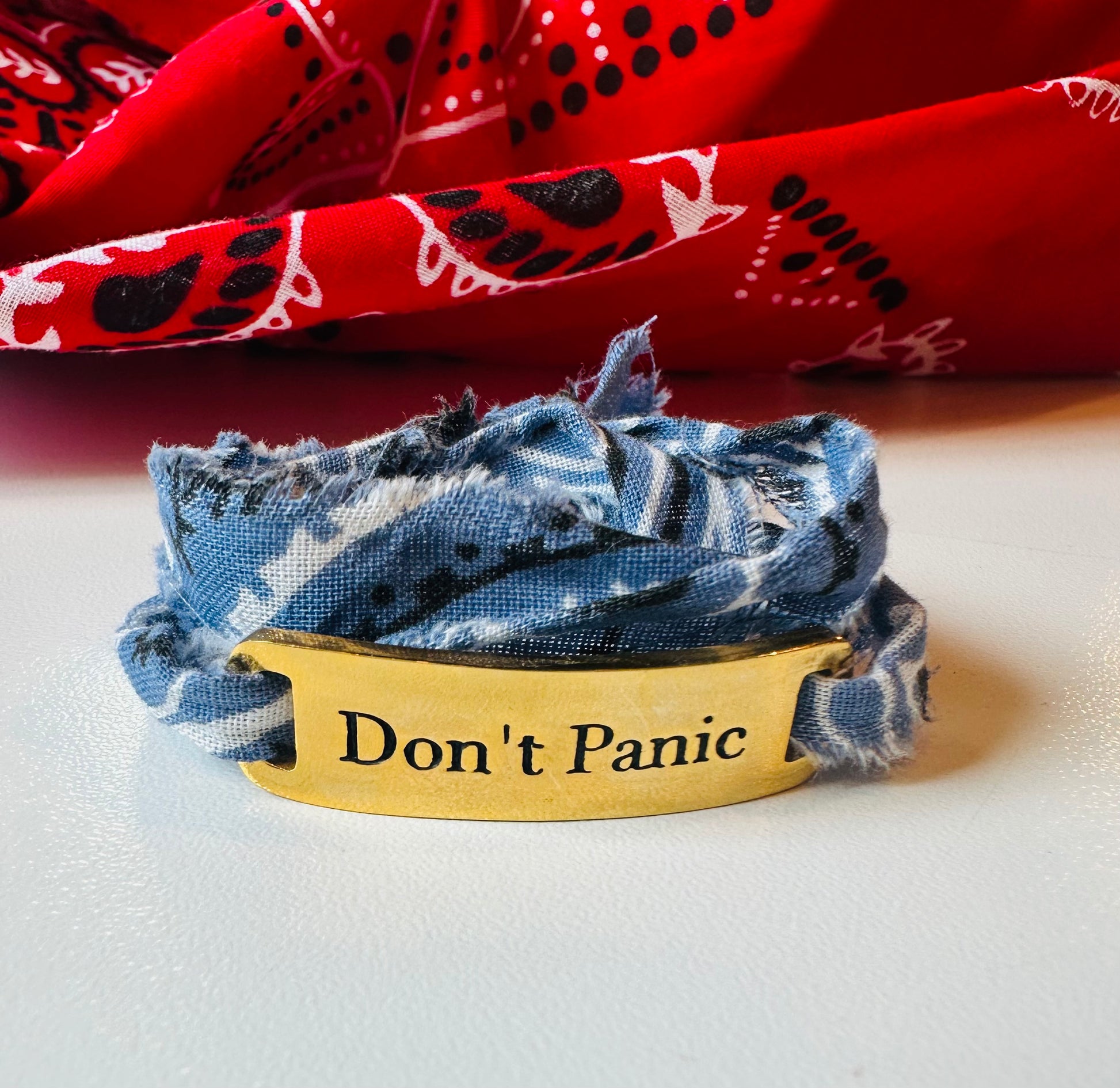 The Simply Eartha Don't Panic Bandana Wrap Bracelet features a limited gold plated design engraved with "Don't Panic," wrapped in frayed blue fabric, set against a vibrant backdrop of red cloth with black and white patterns.