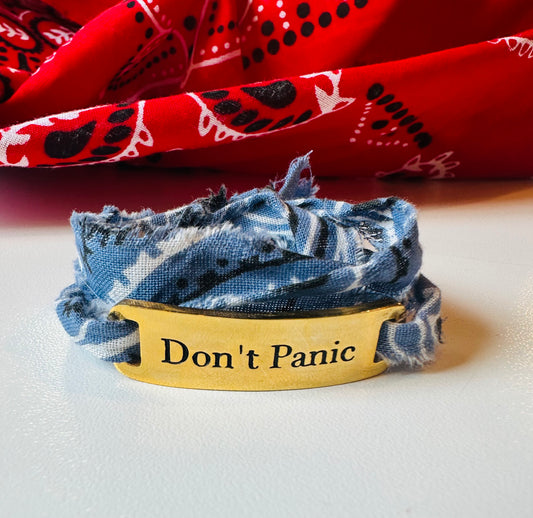 The Simply Eartha Don't Panic Bandana Wrap Bracelet features a limited gold plated design engraved with "Don't Panic," wrapped in frayed blue fabric, set against a vibrant backdrop of red cloth with black and white patterns.