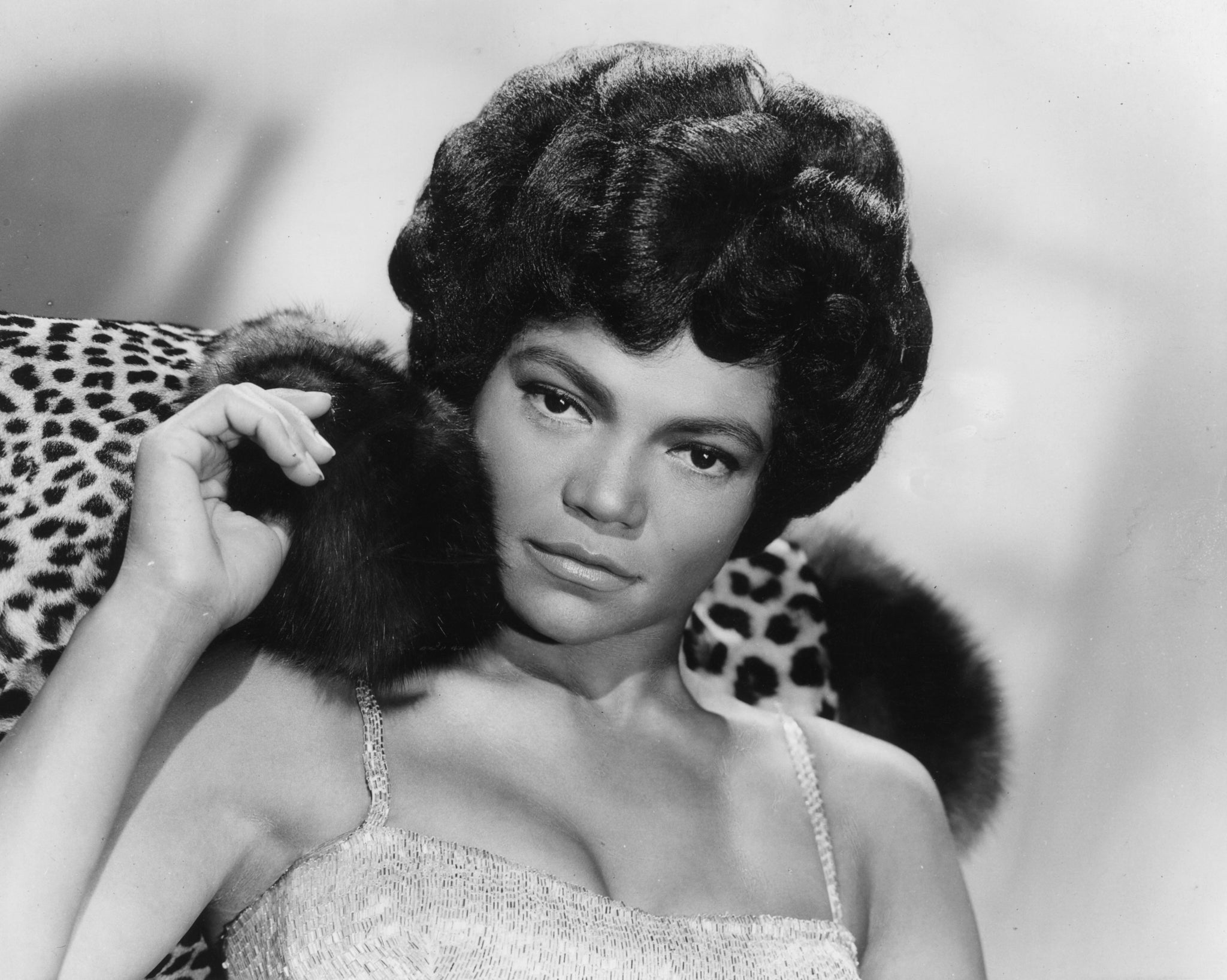 A woman with styled hair, wearing a textured sleeveless top, reclines against a leopard print cushion. She holds a piece of fur delicately in her right hand, gazing directly at the camera with a calm expression. The image is in black and white.