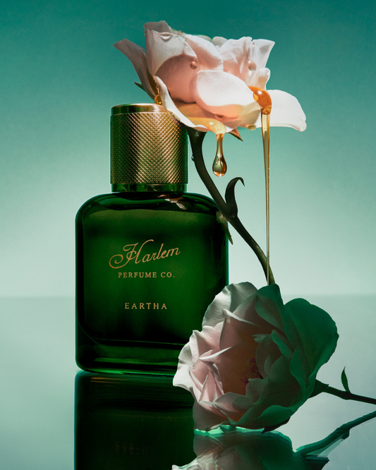 A green bottle of Simply Eartha EARTHA EAU DE PARFUM stands beside two pale pink roses, golden honey dripping from a bloom onto the bottle, all set against a vibrant teal gradient background—an ode to Eartha Kitt.