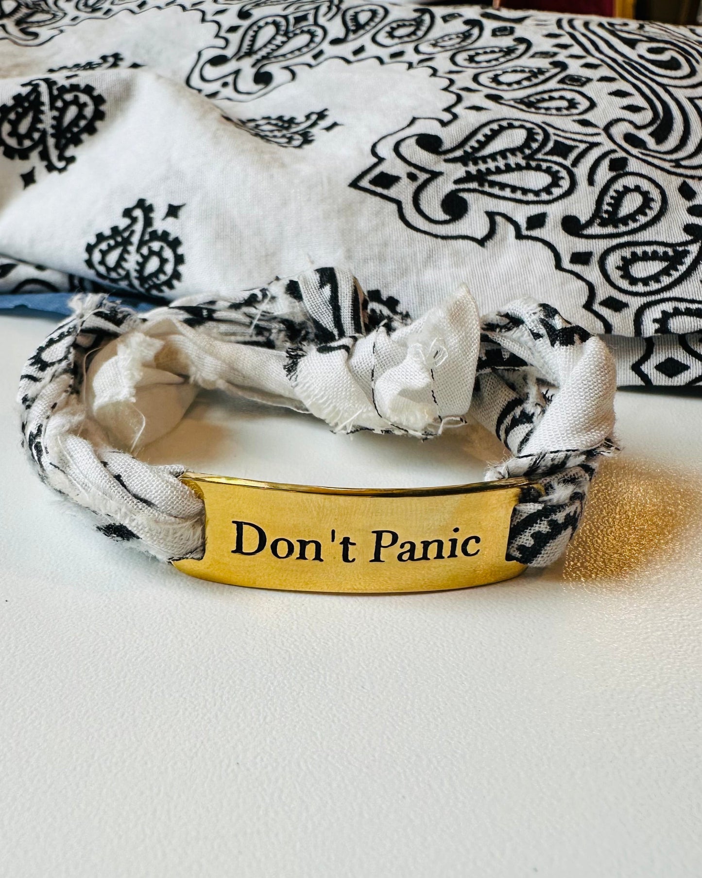 The Simply Eartha Don't Panic Bandana Wrap Bracelet, featuring a gold plated brass plate inscribed "Don't Panic," rests on a white surface with a black and white patterned cloth in the background.